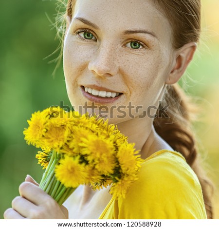 Portrait of red-haired smiling beautiful young woman in yellow blouse with bouquet of dandelions, against green of summer park.