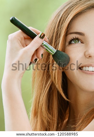 Half face of miling beautiful young woman with bare shoulders on face powder, against green of summer park.