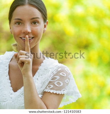 Dark-haired smiling beautiful young woman in white blouse puts finger to his lips, against green of summer park.