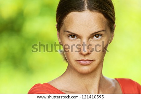 Dark-haired sad beautiful young woman in red blouse, against green of summer park.