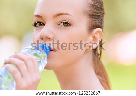 Beautiful young woman drinks water from bottle, against background of summer green park.
