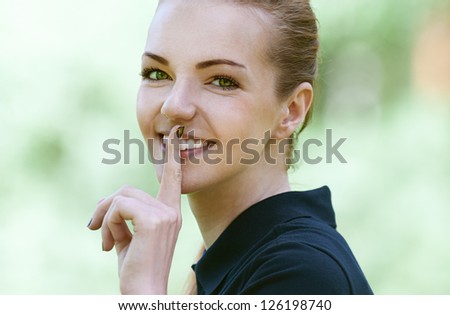 Beautiful smiling young woman in dark blouse, put finger to his lips, against green of summer park.