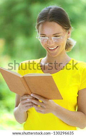 Dark-haired smiling beautiful young woman in yellow blouse and glasses reading books, against green of summer park.