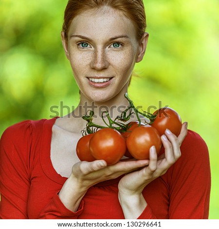 Portrait of red-haired smiling beautiful young woman in red blouse with tomato, against green of summer park.