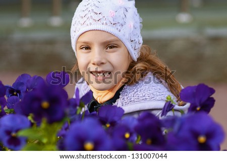 Beautiful funny little girl in jacket and hat, against background of violet flowers.