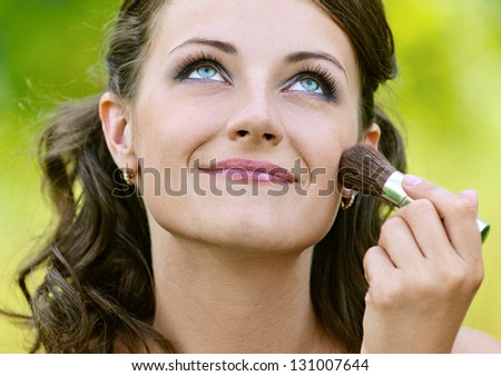 Beautiful young woman close up puts on her face powder brush, against green of summer park.