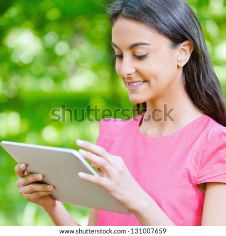 Portrait of beautiful smiling dark-haired young woman in pink dress with e-book, against summer green park.