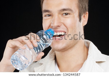 Young handsome man drinks water from bottle, on black background.