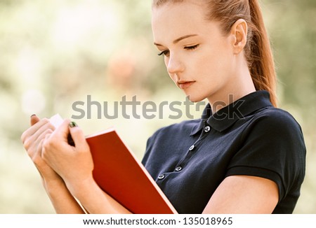Beautiful young woman in dark blouse reads red book, against green of summer park.