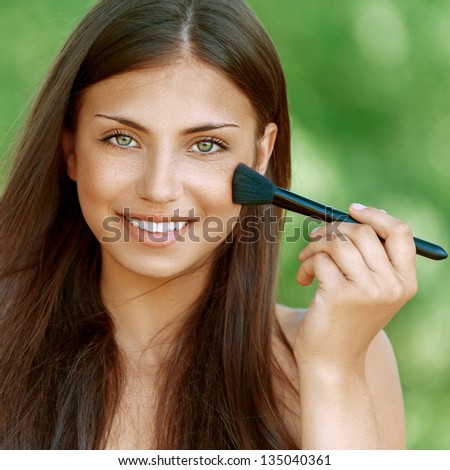 Beautiful dark-haired smiling young woman treats her face with brush, against background of summer green park.