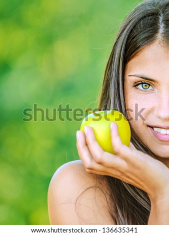 Portrait half of face young beautiful woman with bare shoulders holding an apple and smiling, on green background summer nature.