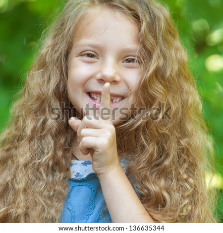 Pretty smiling little girl puts finger to lips, on green summer background.