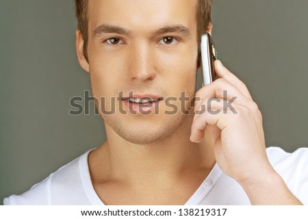 Smiling young man in white shirt talking on cell phone, on grey background.