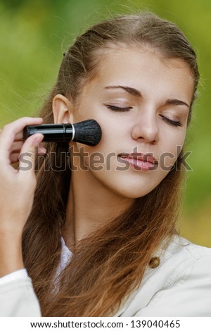 Young beautiful woman with closed eyes causes powder to face, against background of autumn park.