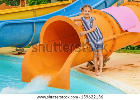 Beautiful little girl stands near large orange water park slides.