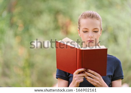 Beautiful young woman in dark blouse reads red book, against green of summer park.