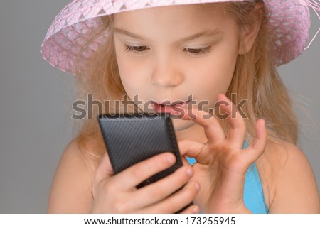 Little smiling girl in red dress reading sms on your cell phone, on grey background.