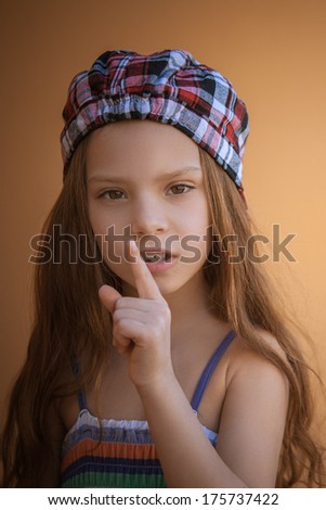 Beautiful sad little girl puts index finger to lips, on yellow background.