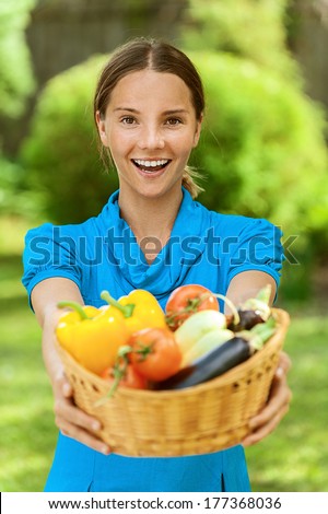 Portrait of dark-haired smiling beautiful young woman in blue blouse with baskets of fruit and vegetables, against green of summer park.