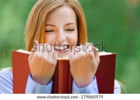 Beautiful smiling young woman reading red book, against background of summer green park.