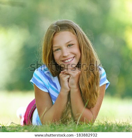 Beautiful smiling teenage girl in blue blouse lying on grass, against green of summer park.