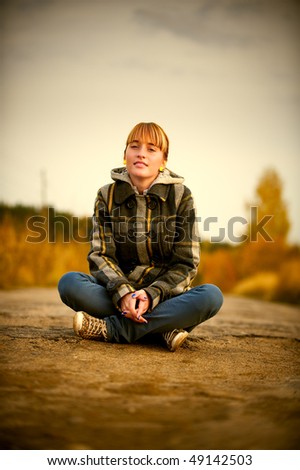 Charming young woman sits in middle of road against autumn nature.