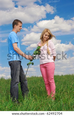 young couple outdoor on green grass. guy granting flower, girl looking shy. blue sky with clouds on background.