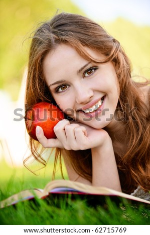 Charming girl with apple lies on green grass and reads book.