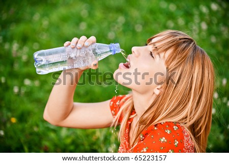 Beautiful girl in red dress drinks water from bottle, against green grass.