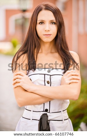 Sad young woman in white dress with long hair and crossed hands, against city landscape.