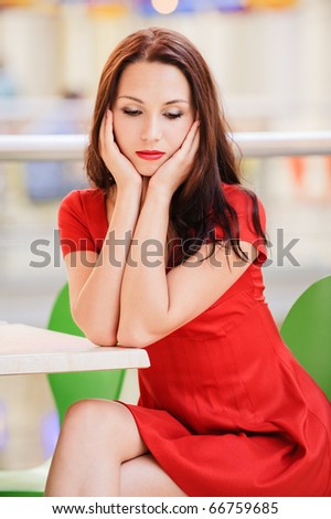Young beautiful dark-haired woman in red dress sits at little table in small cafe and thinks of unfortunate life.