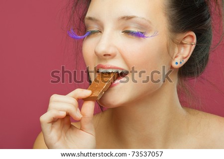 Beautiful young woman has closed eyes and bites off from chocolate brick, on red background.