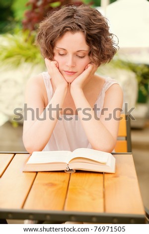 outside portrait of beautiful curly young woman sitting at the park cafe table and reading book