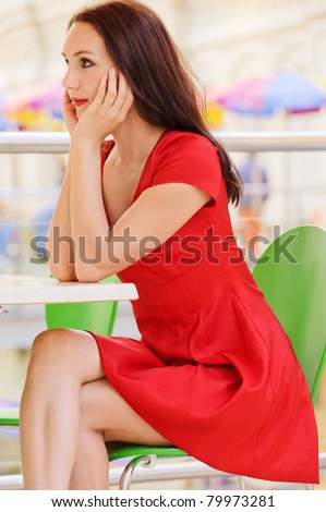 Beautiful young woman in red dress sits at little table in cafe and order expects.