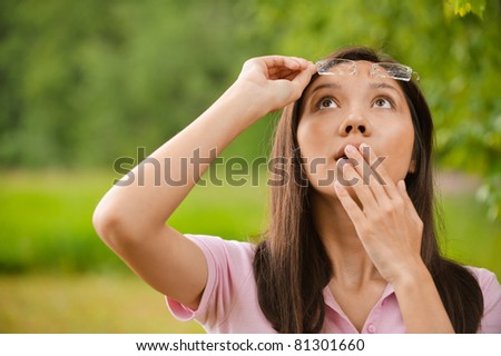 Portrait of beautiful young brunette frustrated woman with eyeglasses staring at sky and wondering in green summer park.