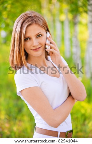 portrait of young beautiful blond woman in white blouse speaking on mobile phone and standing at park