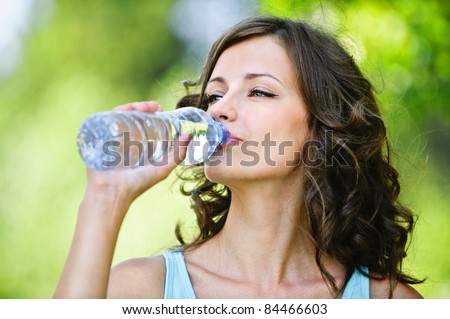 Portrait of young beautiful dark-haired woman wearing blue t-shirt drinking water at summer green park.