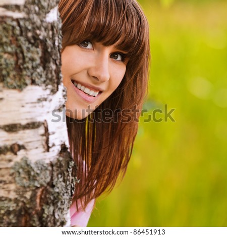 Portrait of young attractive funny smiling dark-haired girl looking from behind birch tree at summer green park.