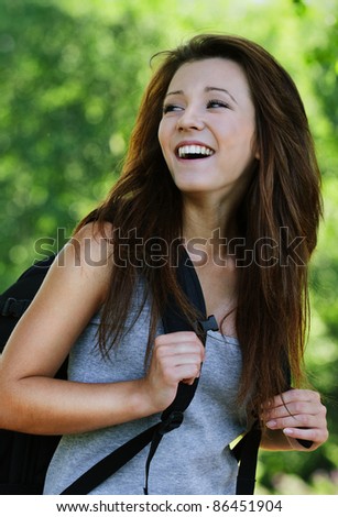Portrait of positive laughing dark-haired girl looking back, wearing grey t-shirt and black backpack at summer green park.