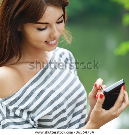 Portrait of young charming smiling brunette woman wearing striped blouse working with stylus and smartphone at summer green park.