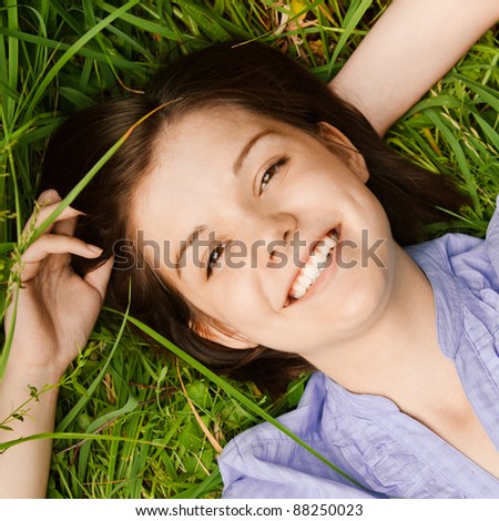 Portrait of young cute dark-haired smiling woman wearing blue blouse lying on grass at summer green park.