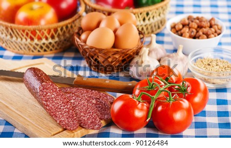 foods (tomatoes, eggs, apples, Bulgarian pepper, hazelnuts, garlic and smoked sausage on cutting board with knife) on blue and white checkered tablecloth