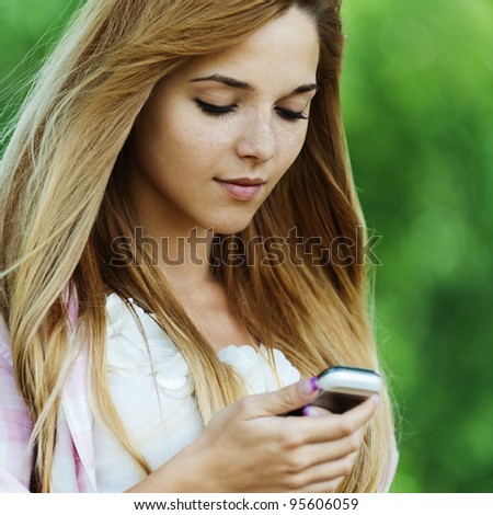 Portrait of young pretty, long-haired, sad woman standing against background of summer green of park, holding mobile phone