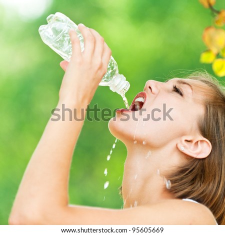 portrait of young woman in profile with short hair quenches thirst (drinks from bottle of water), against backdrop of summer nature