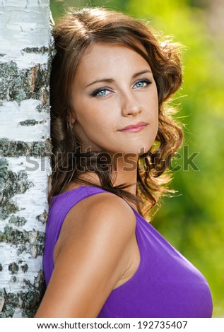 Beautiful young woman in purple dress is based on sad birch, against green of summer park.