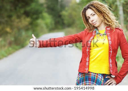 Young beautiful woman with dreadlocks in red clothes votes hitchhiking on road.
