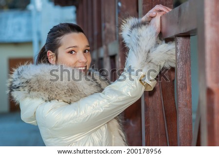 Young smiling beautiful woman in white coat with fur collar around wooden railing.