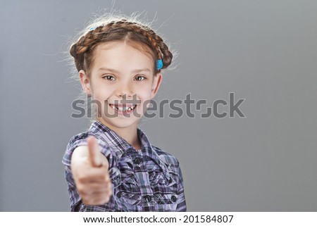 Little cheerful girl lifts thumb upwards, on gray background.