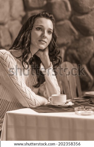 Young beautiful woman sits in cozy cafe, looks out of window and drinks hot tea from cup.