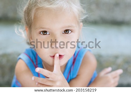 Beautiful little blonde girl with curly hair puts her finger to mouth, against background of summer city park.